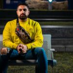 A man in a yellow sweater and jeans sits on an outdoor bench at night.