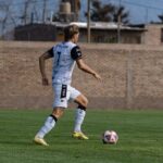 Soccer player dribbles the ball on a grassy field against a residential backdrop. Action captured during the day.