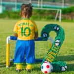 A young child wearing a Neymar Jr. jersey stands by a mini soccer goal on a grassy field.