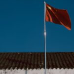 Chinese flag waving over traditional building roof against a clear blue sky.