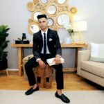 A fashionable young man in a suit sitting in a stylish, modern living room with wooden decor.