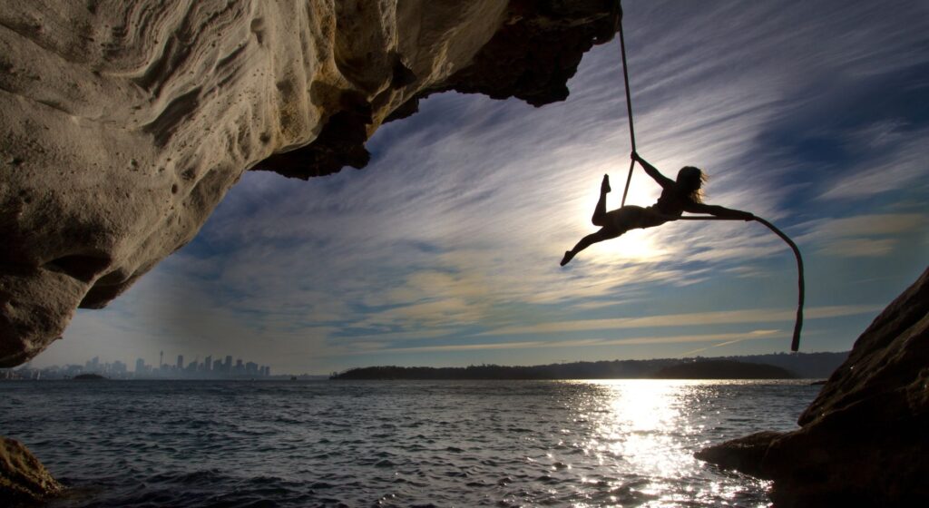 Silhouette of a woman performing acrobatics on a rope hanging from a cliff by the sea with city skyline in the background.