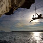 Silhouette of a woman performing acrobatics on a rope hanging from a cliff by the sea with city skyline in the background.