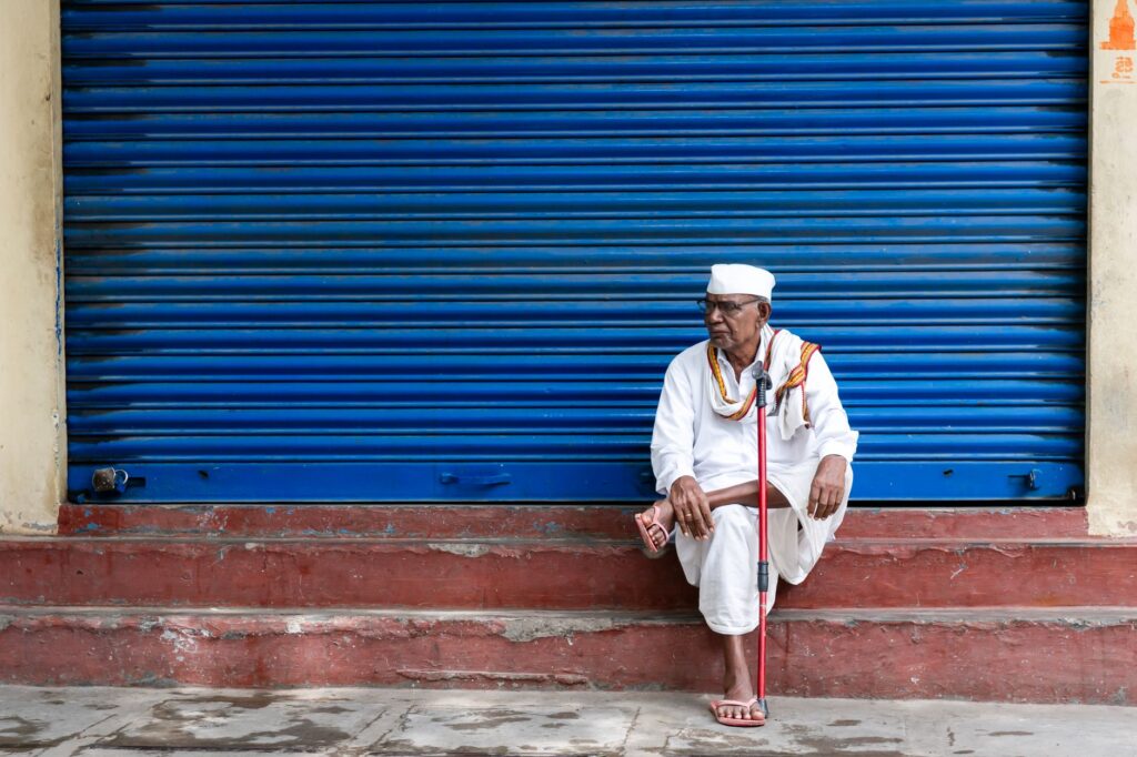 Elderly man in traditional attire sitting with cane by closed blue shutters.
