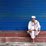 Elderly man in traditional attire sitting with cane by closed blue shutters.