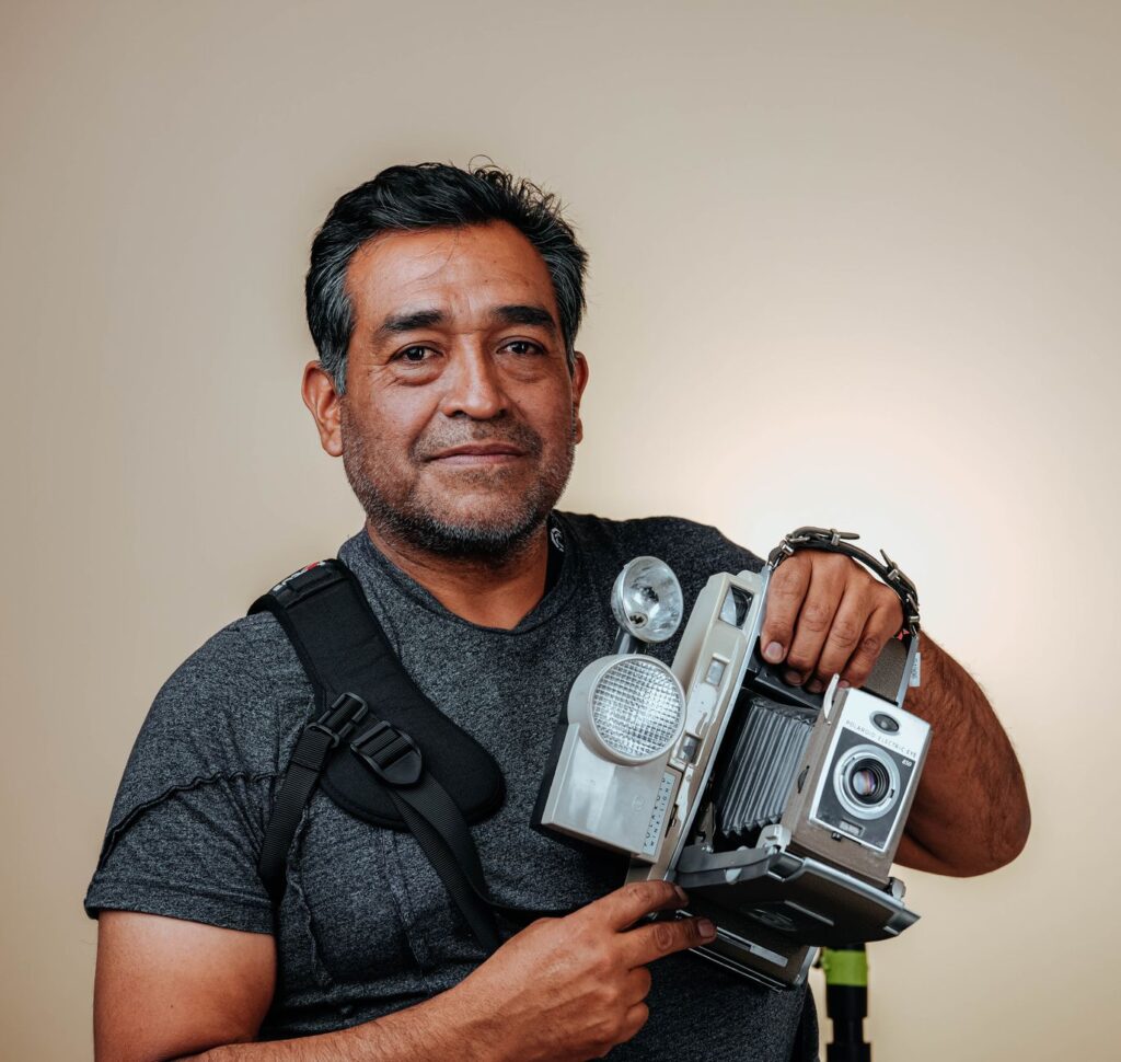 Vintage-style portrait of a man holding an old-fashioned camera indoors with a beige backdrop.