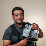 Vintage-style portrait of a man holding an old-fashioned camera indoors with a beige backdrop.