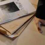 Close-up of a hand resting on a desk near a stack of investigative documents and newspaper clippings.