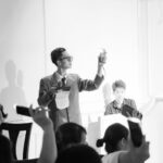 Elegant black and white photo of an auction scene with a presenter and bidders raising phones.