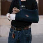 A man wearing a dark sling with a bandaged arm, standing outdoors in casual attire.