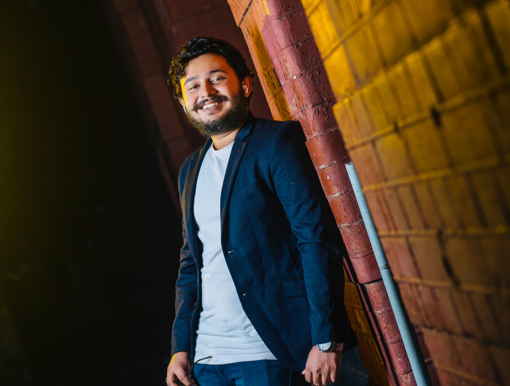 A bearded man in a blazer smiles as he leans against a brick wall. Captured in Sri Lanka.