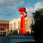 Striking red modern sculpture in a cityscape with historic buildings under a blue sky.