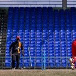 A cricket player in red batting during a match in an empty stadium with blue seats.