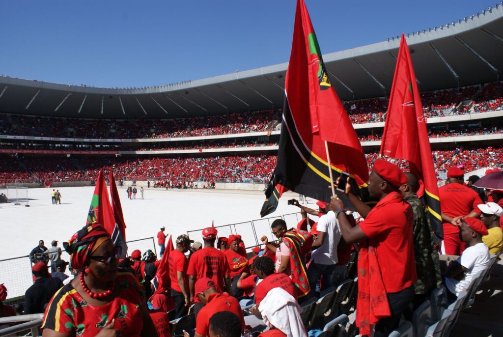 Colorful crowd in a stadium in Soweto, South Africa, waving red flags and wearing red attire.