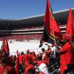 Colorful crowd in a stadium in Soweto, South Africa, waving red flags and wearing red attire.