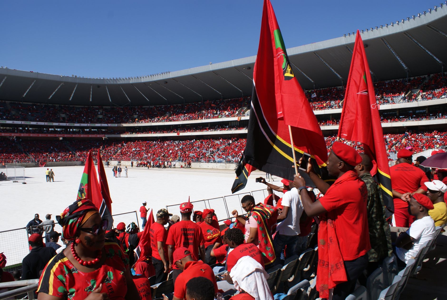 Colorful crowd in a stadium in Soweto, South Africa, waving red flags and wearing red attire.
