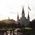 St. Louis Cathedral in New Orleans with an American flag, trees, and a garden at dusk. Iconic religious landmark.