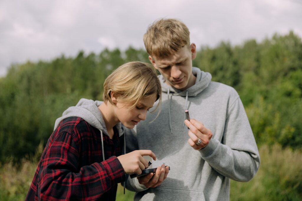 Two teenagers focus intently on a device outdoors, surrounded by nature.