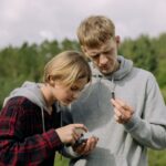 Two teenagers focus intently on a device outdoors, surrounded by nature.