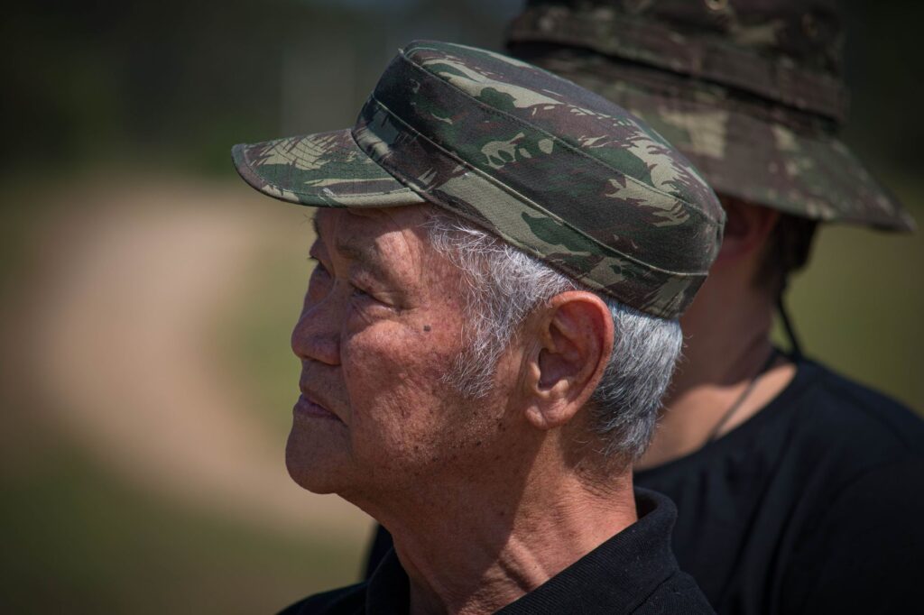 Elderly Asian man wearing a camouflage military cap, outdoors.