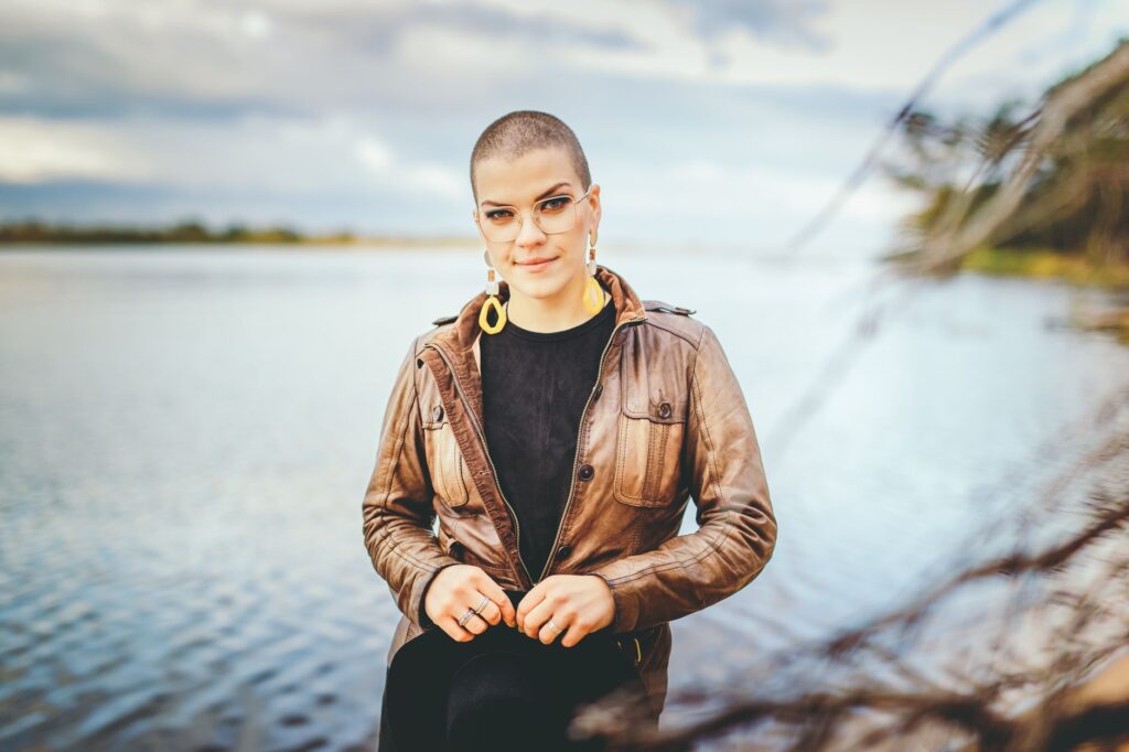 A stylish woman with short hair and glasses poses by a scenic body of water.