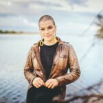 A stylish woman with short hair and glasses poses by a scenic body of water.