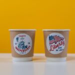 Two coffee cups with Independence Day designs on a yellow background.