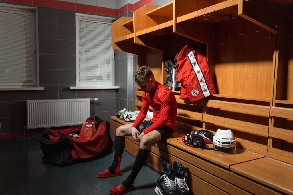 Hockey player in red sportswear sitting on a wooden bench in a locker room, preparing for a game.