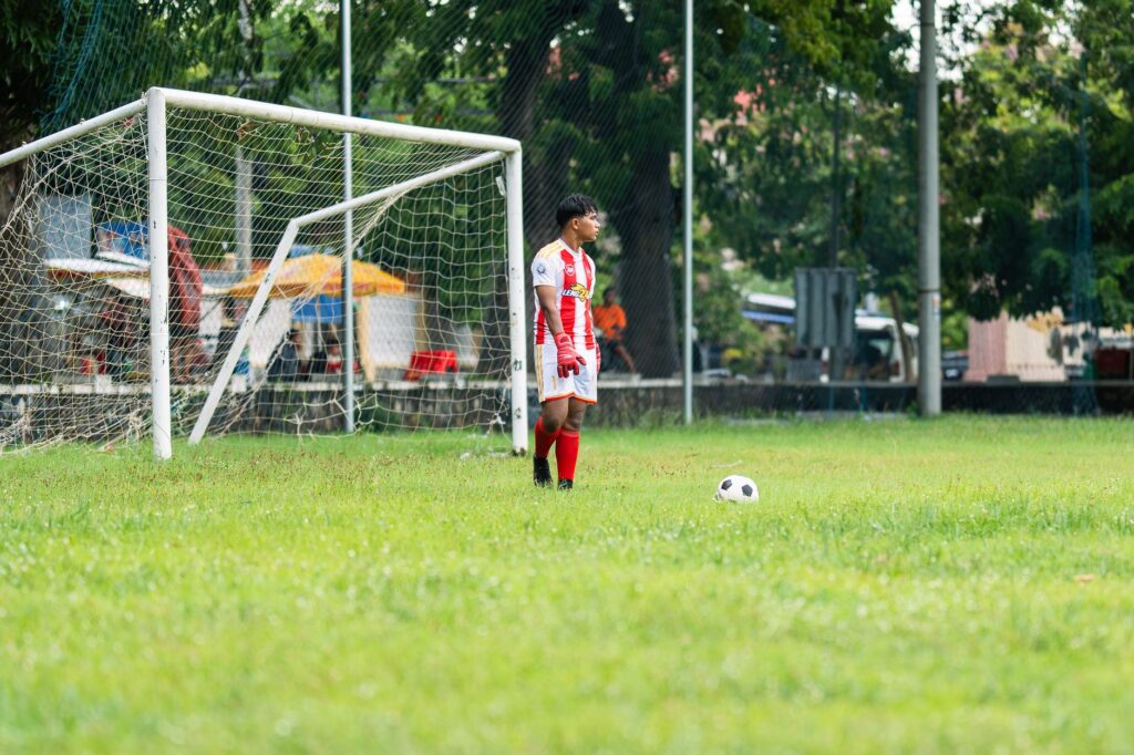 Soccer goalkeeper in action on outdoor field. Player in red kit ready near goal.