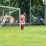 Soccer goalkeeper in action on outdoor field. Player in red kit ready near goal.