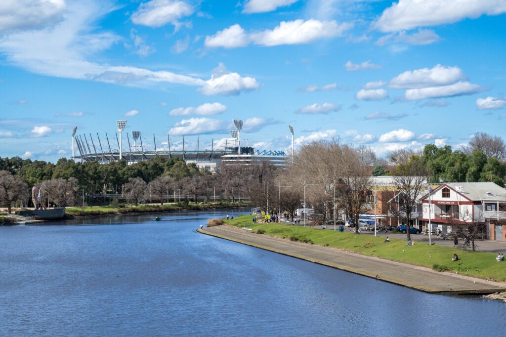 A scenic view of the Melbourne Cricket Ground alongside the Yarra River under a bright blue sky.