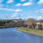 A scenic view of the Melbourne Cricket Ground alongside the Yarra River under a bright blue sky.