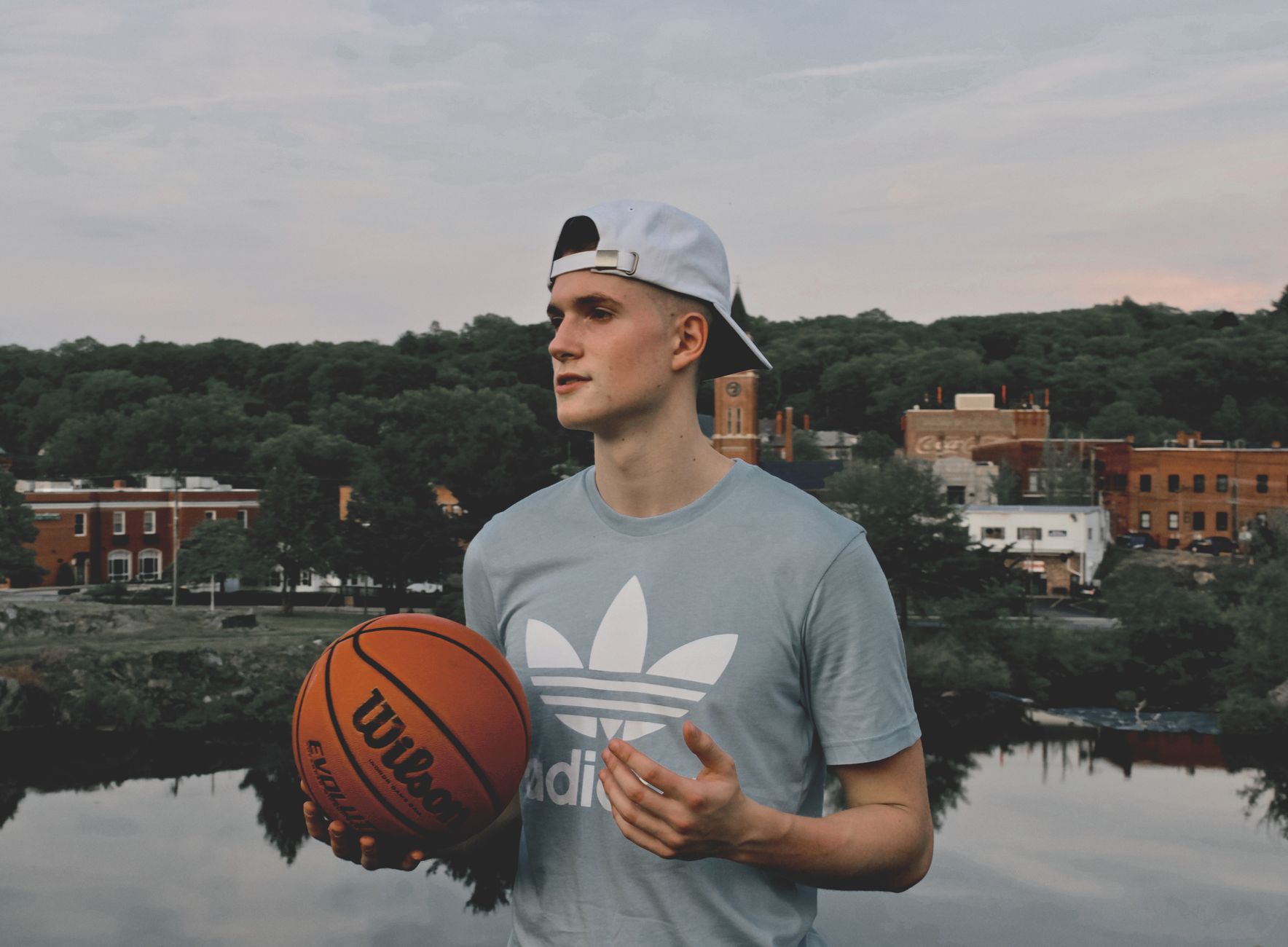 Young man holding a basketball by a lake in Putnam, CT, with town buildings in the background.