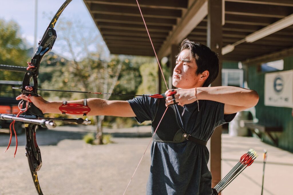 Asian man practicing archery with a recurve bow outdoors on a sunny day, focused and aiming.