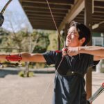 Asian man practicing archery with a recurve bow outdoors on a sunny day, focused and aiming.