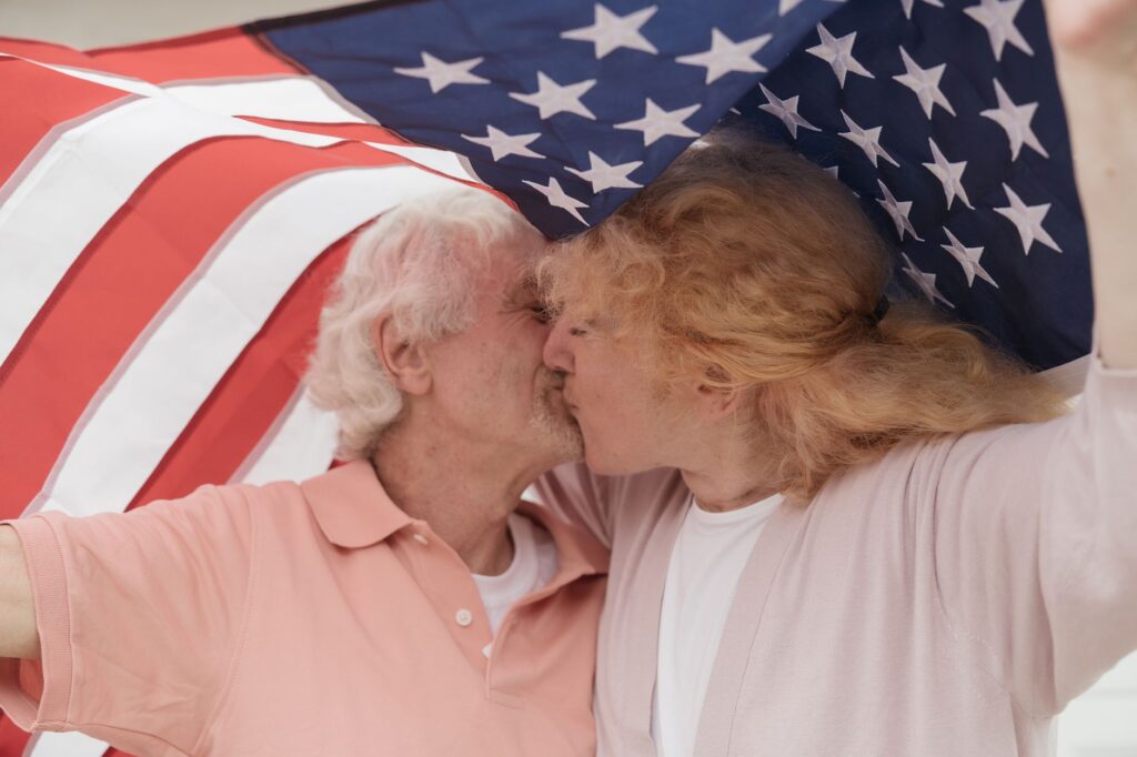 Senior couple sharing a kiss under an American flag, symbolizing love and patriotism.