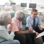 Business professional consults elderly clients in an office setting. Collaborative discussion, paperwork visible.