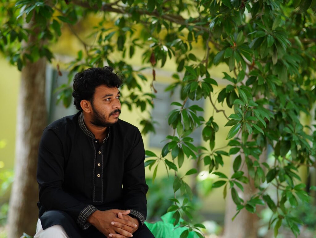 A man in traditional attire sits thoughtfully under a tree outdoors in Dhaka, Bangladesh.